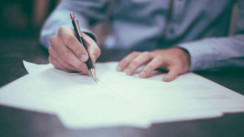 Photo of persons hands on desk, one hand holding a pen and signing paperwork